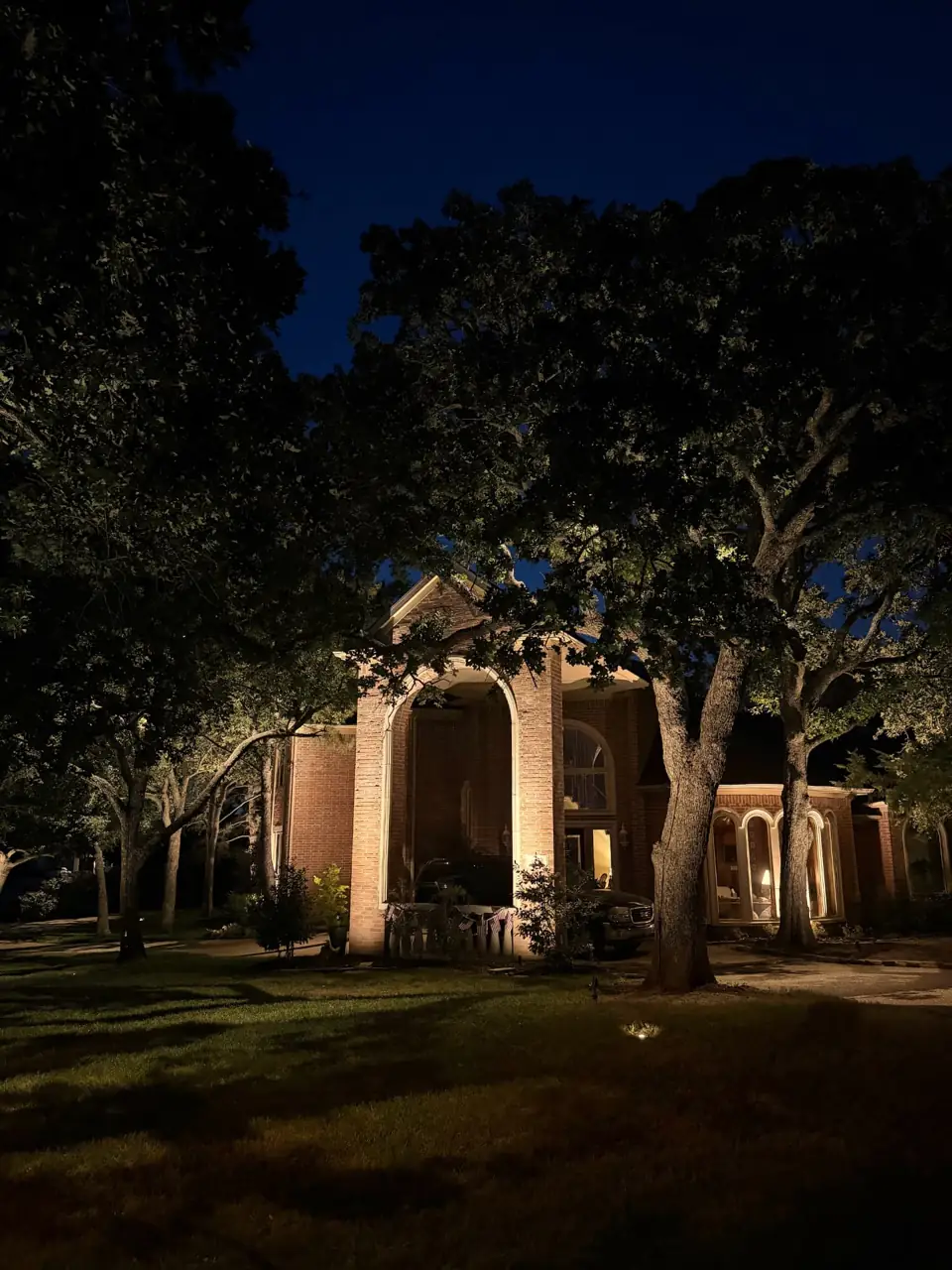 Brick two-story home with arched entry under oak trees after lighting installation